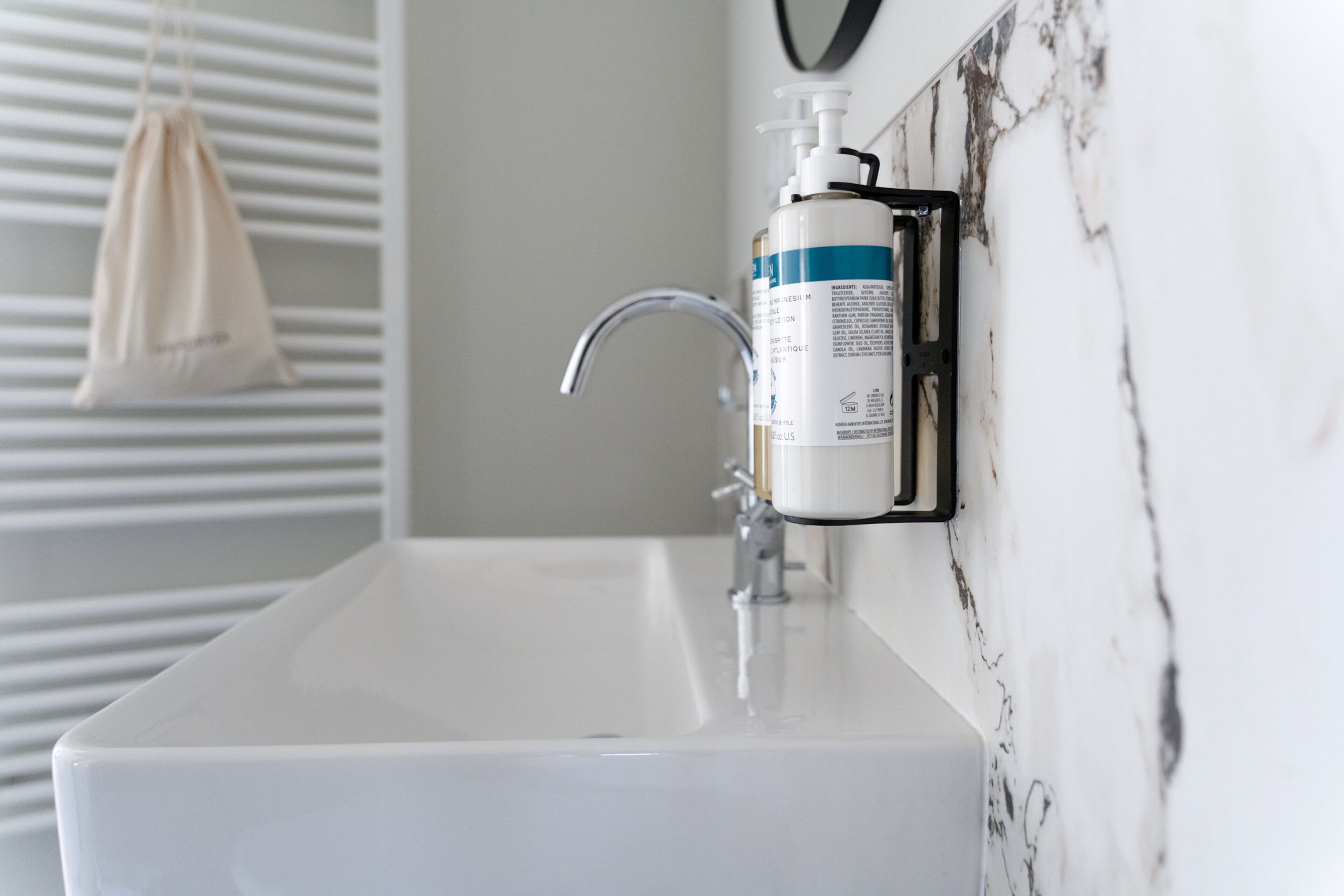 A bathroom with a white sink, a marble wall, and a bottle of hand soap mounted on the wall.