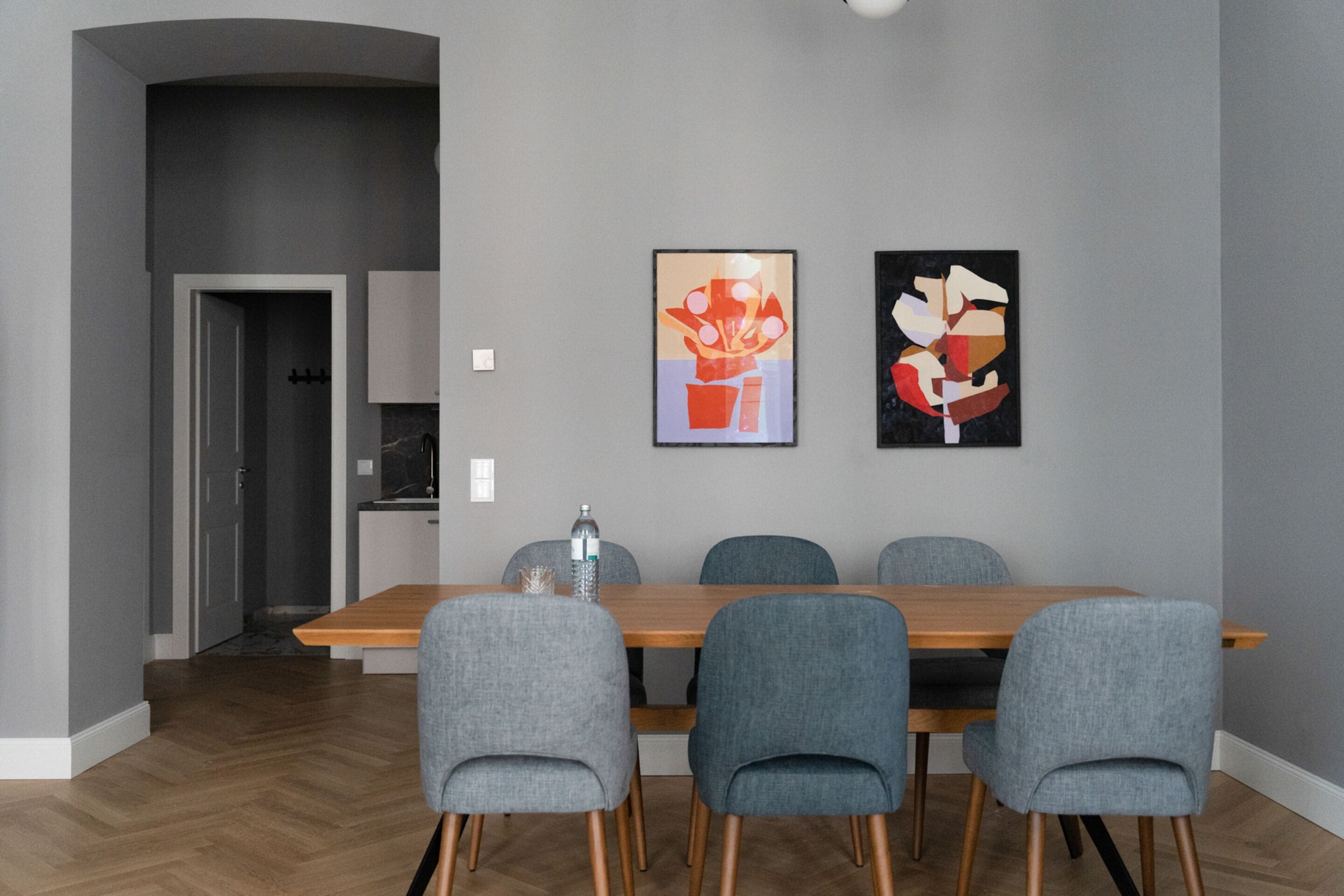 A modern dining area with a wooden table and four grey upholstered chairs. There are two abstract paintings on the wall.