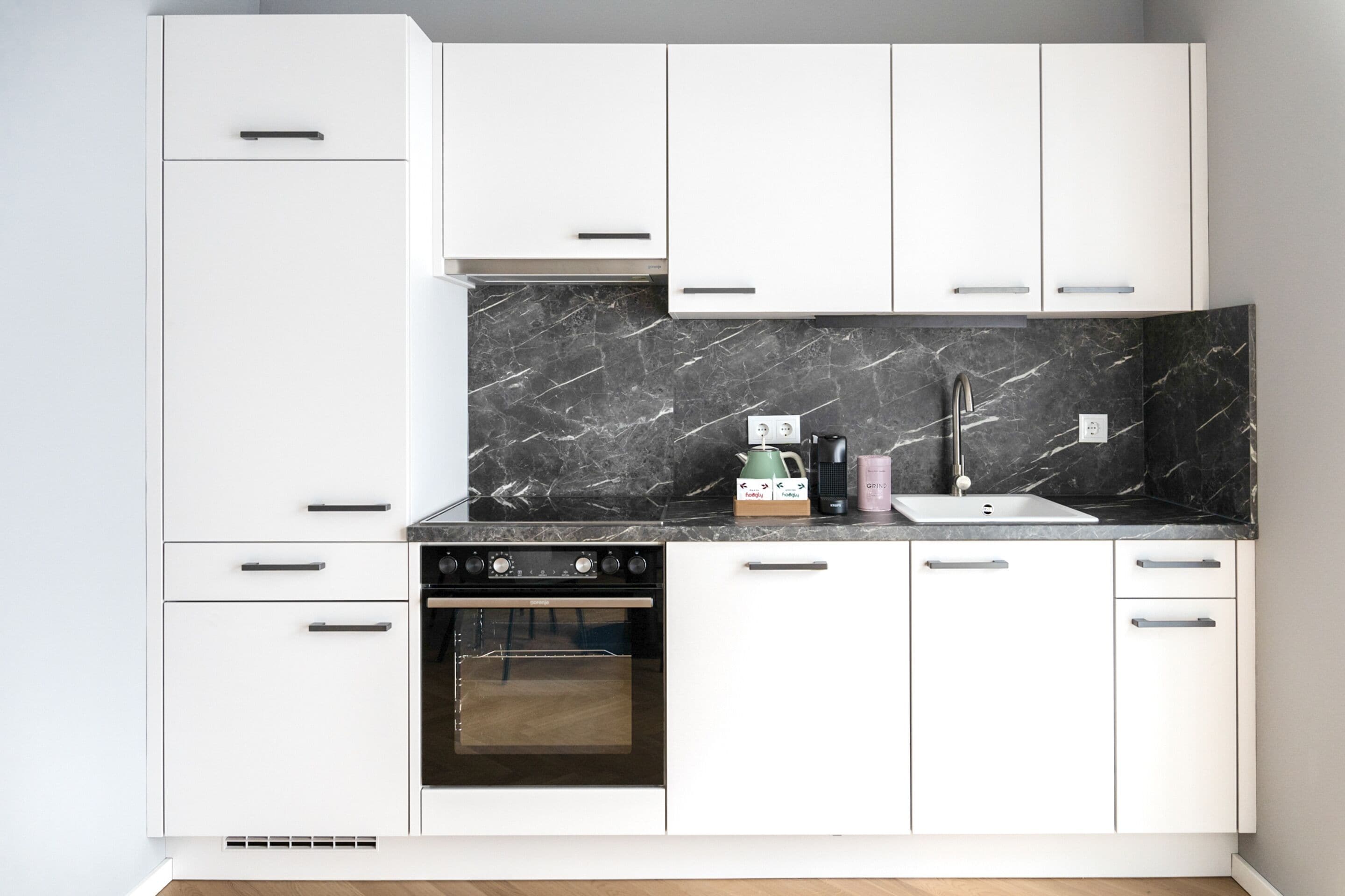 Een moderne keuken met witte kastjes, een zwarte marmeren spatwand, een ingebouwde oven en een roestvrijstalen kraan.