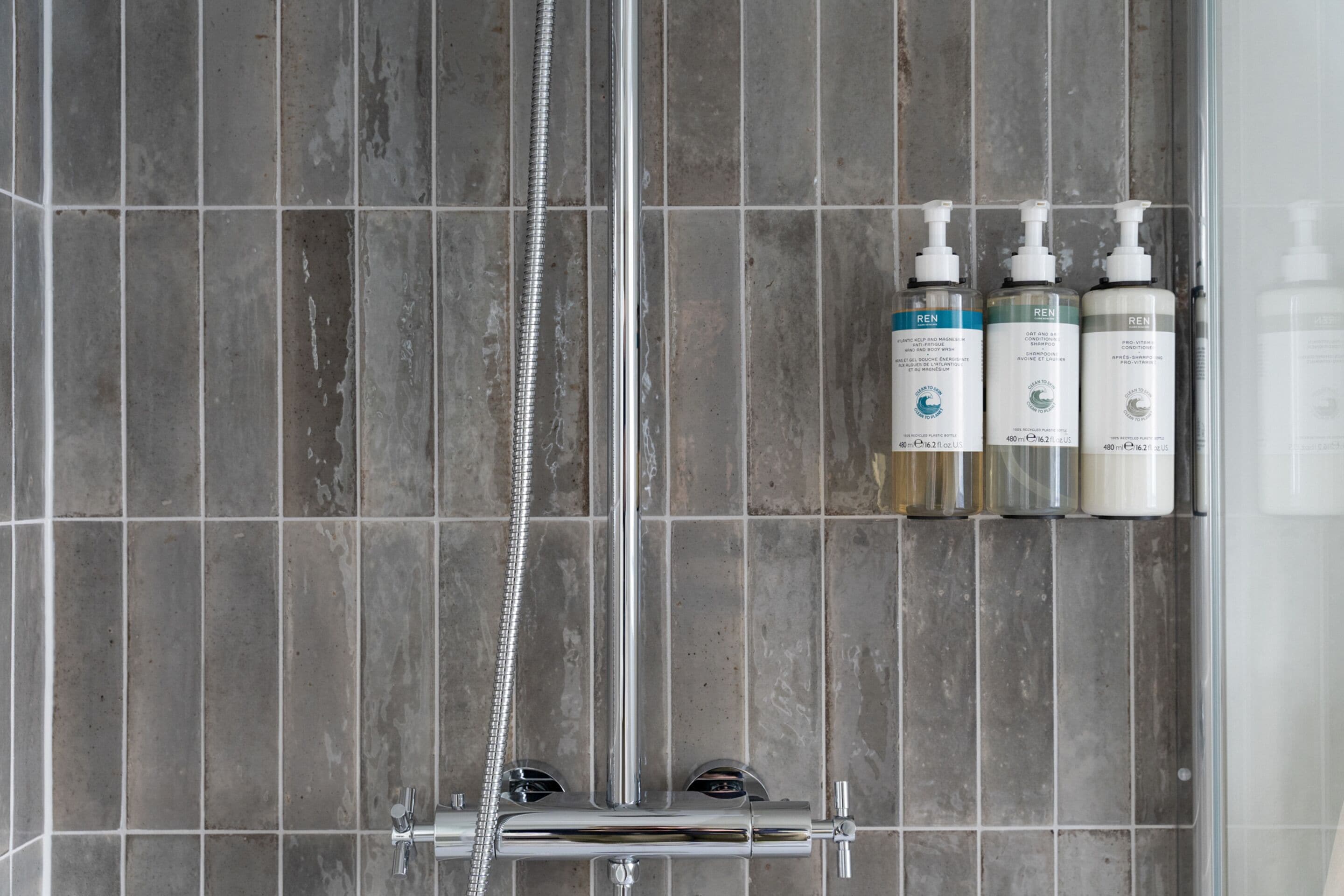 A shower area with grey tiles, a chrome showerhead, and three bottles of Ren shampoo on a shelf.