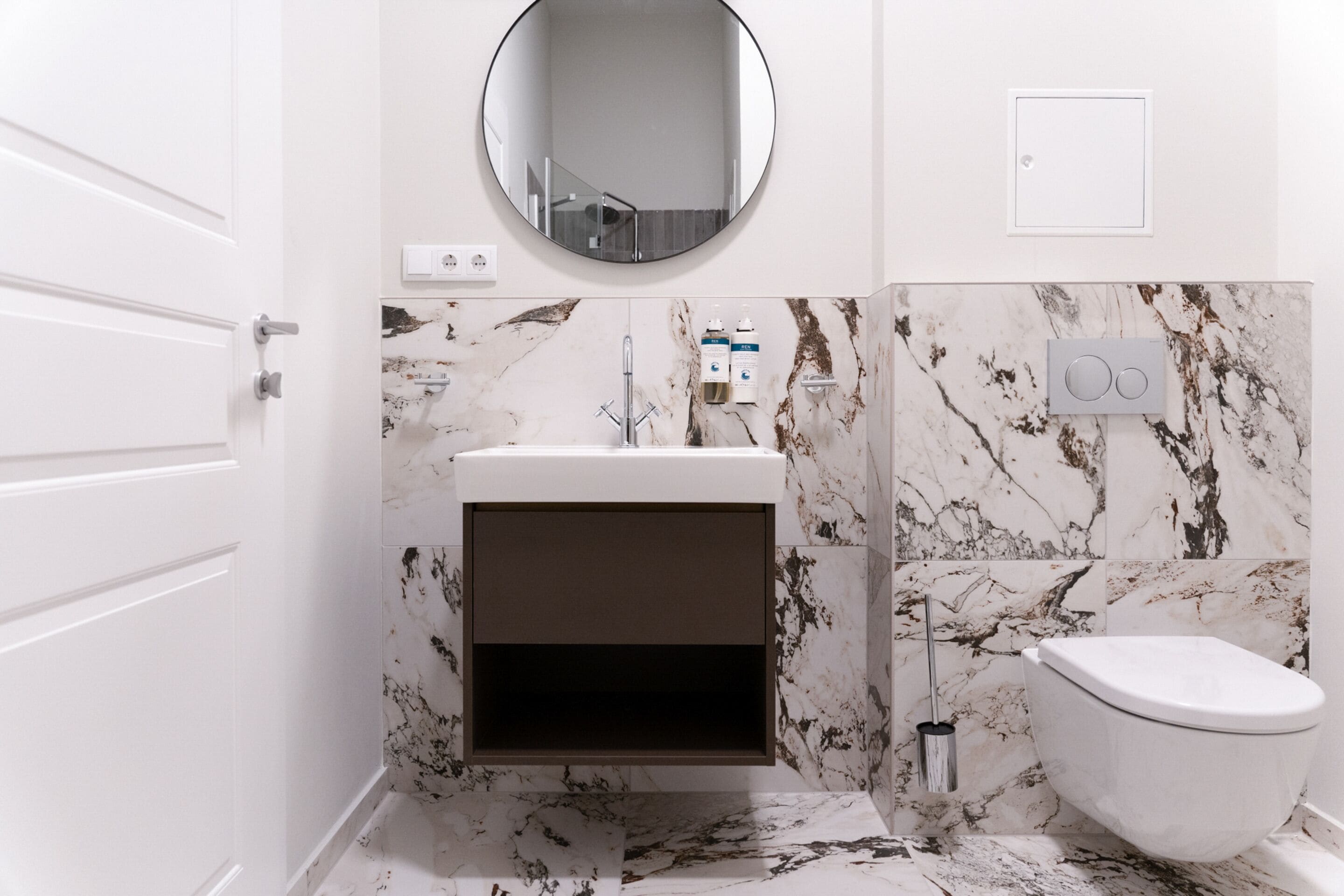 A bathroom with a white vanity, a toilet, a round mirror, and marble-patterned walls.