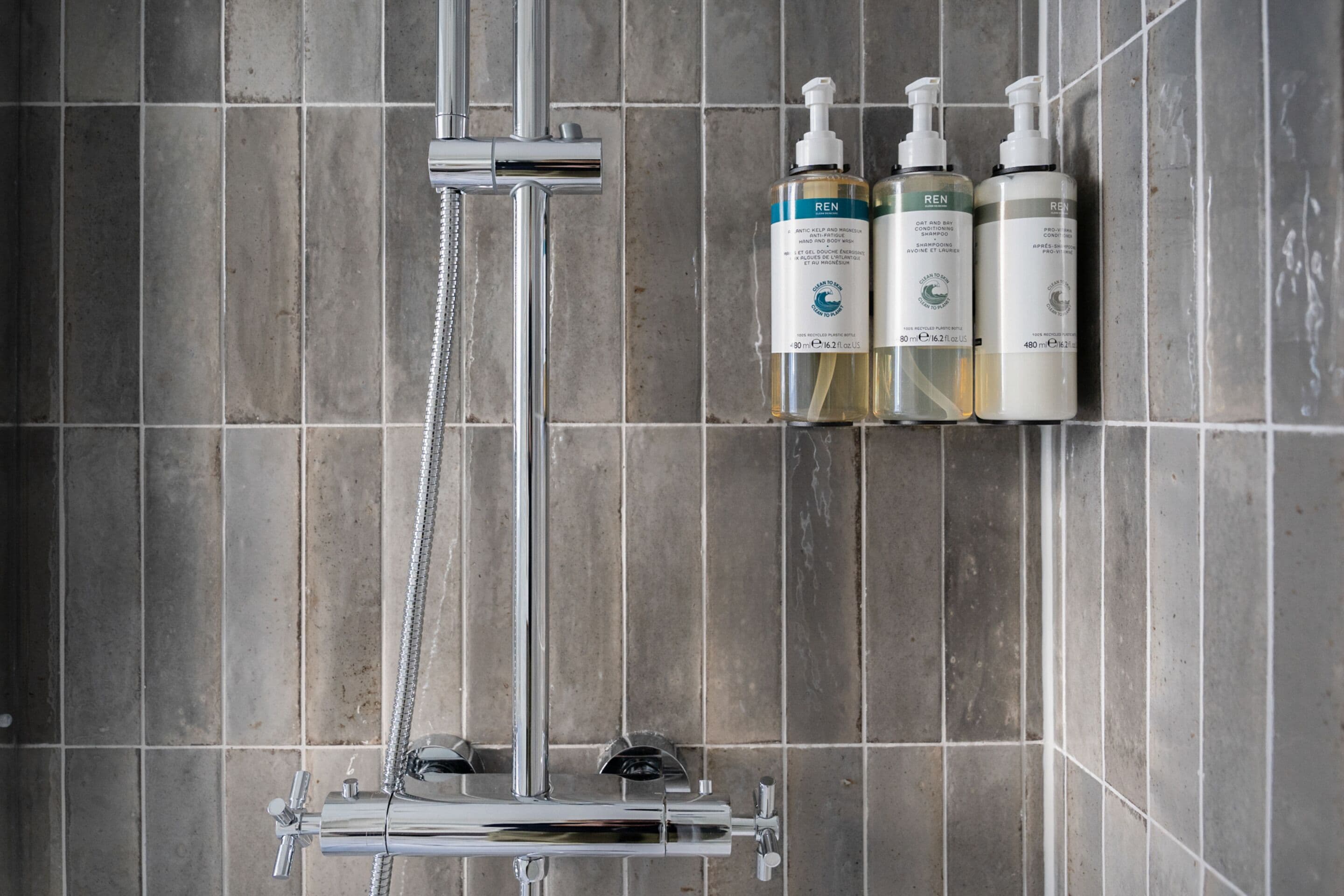 A shower with a chrome faucet and a handheld showerhead. Three bottles of REN shampoo and conditioner are mounted on the wall.