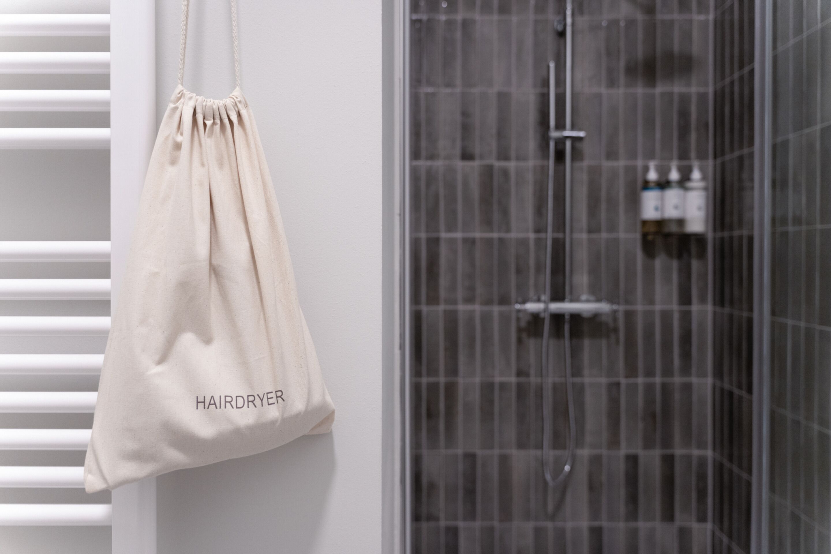 A bathroom with a shower, a hanging beige bag labeled "HAIRDRYER", and a white towel rack.