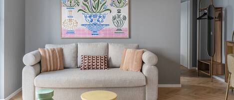 Ein Wohnzimmer mit grauem Sofa, gemusterten Kissen, einem grün-gelben Hocker und einem floralen Kunstwerk an der Wand.
