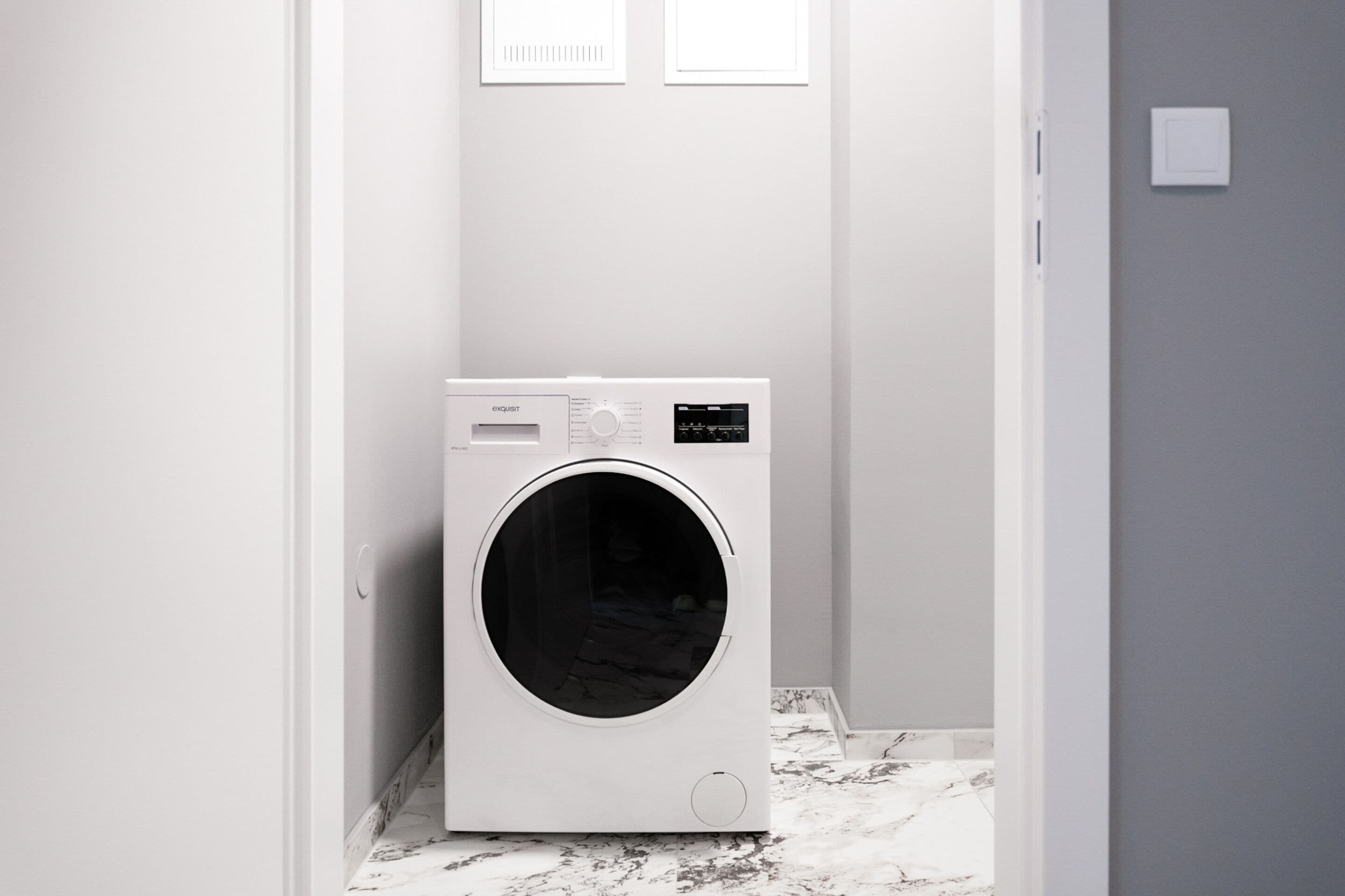 A compact, modern laundry room with a front-loading washing machine, a small control panel, and a wall-mounted vent.