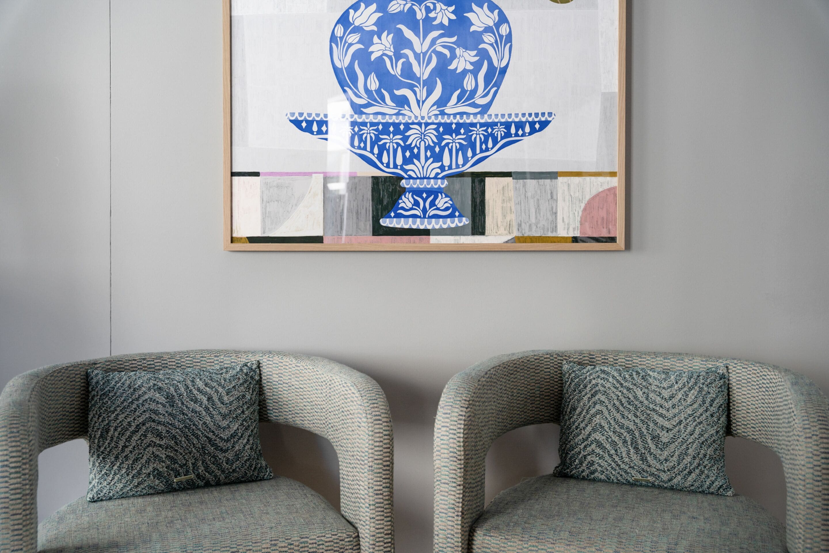 Un'opera d'arte incorniciata con un motivo blu e bianco su una parete, due poltrone grigie con cuscini decorati e un pavimento in legno.