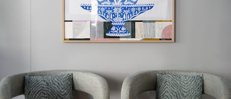 A framed artwork of a blue and white design on a wall, two grey upholstered chairs with patterned cushions, and a wooden floor.