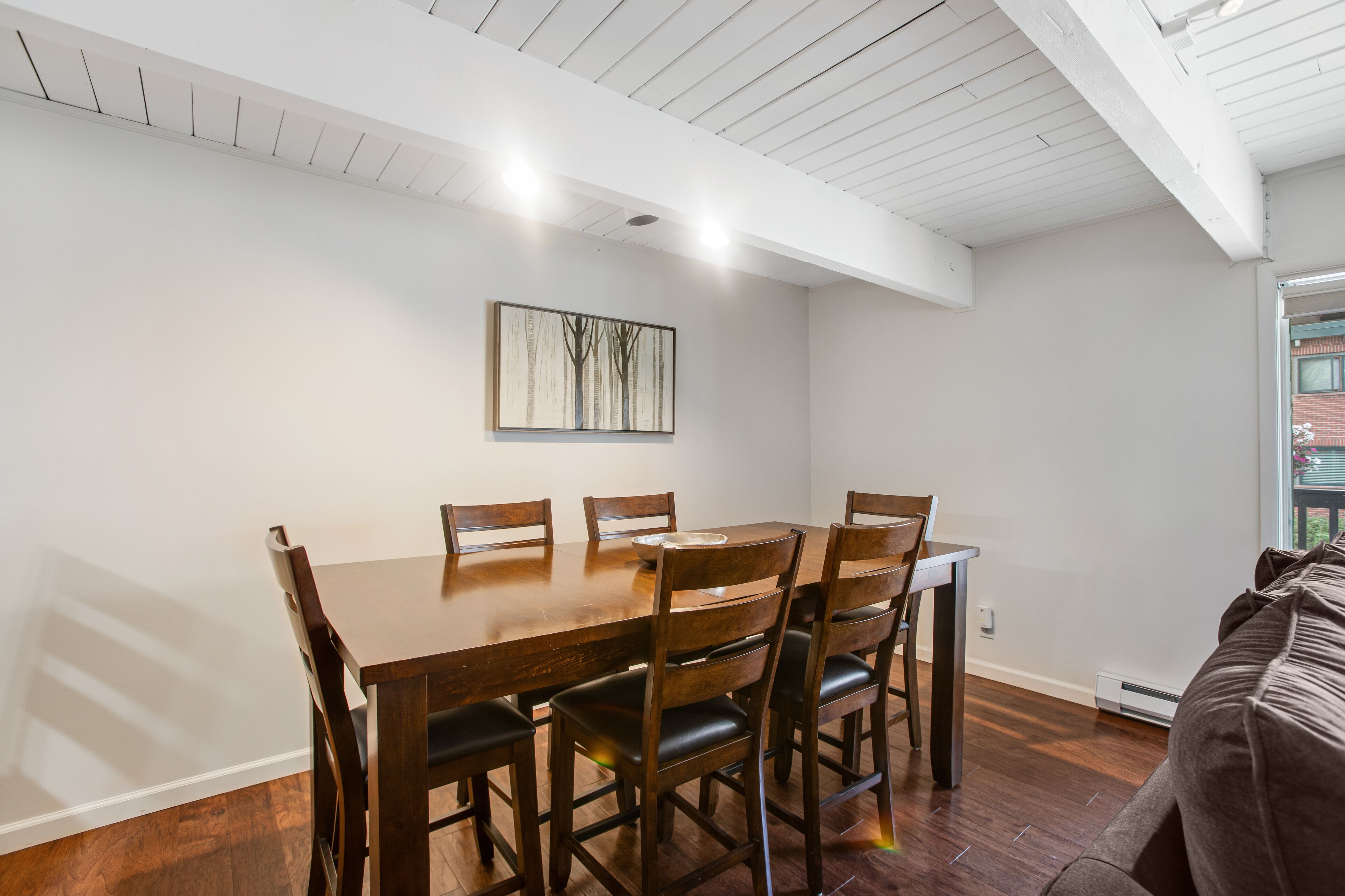 A dining area with a wooden table and chairs, a painting on the wall, and a view of the outdoors through a window.