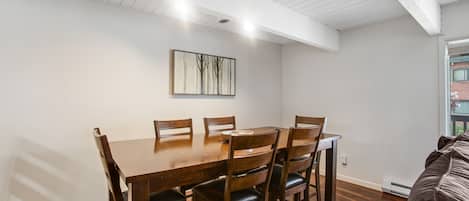 A dining area with a wooden table and chairs, a painting on the wall, and a view of the outdoors through a window.