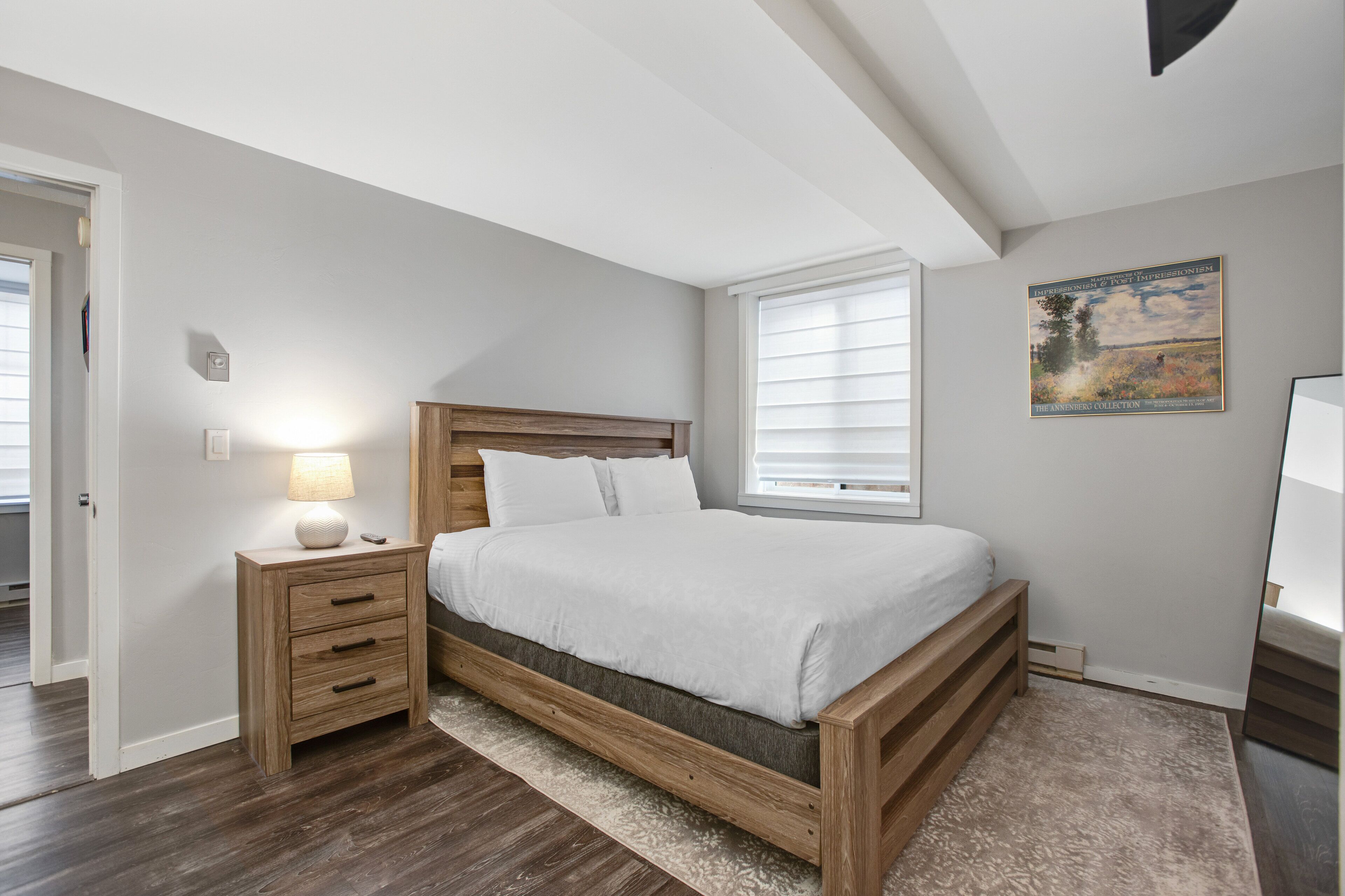 A bedroom with a wooden bed, a nightstand with a lamp, a window with blinds, and a painting on the wall.