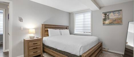 A bedroom with a wooden bed, a nightstand with a lamp, a window with blinds, and a painting on the wall.
