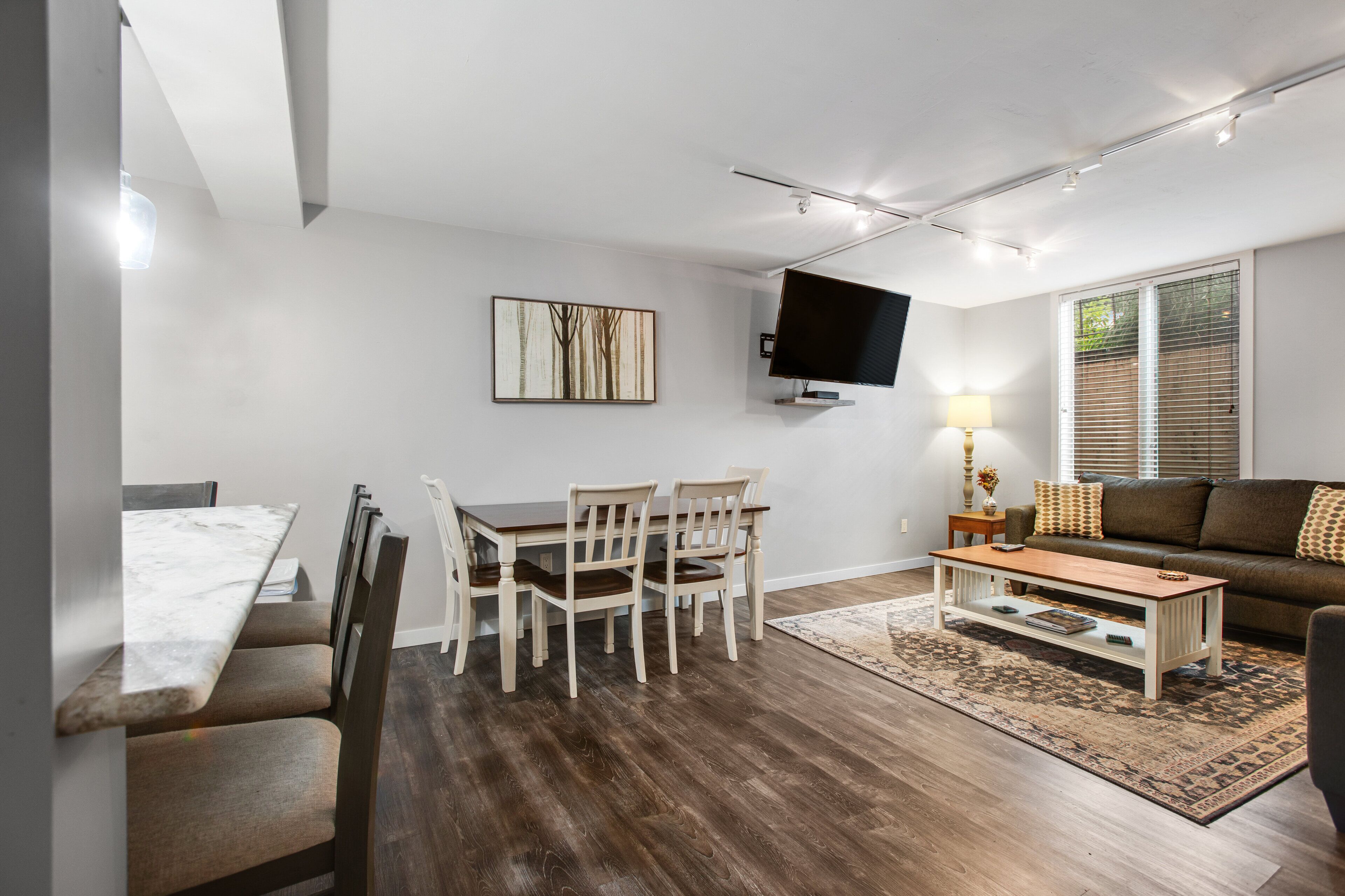 A modern living room with a dining area, a flat-screen TV mounted on the wall, and a large window with blinds.