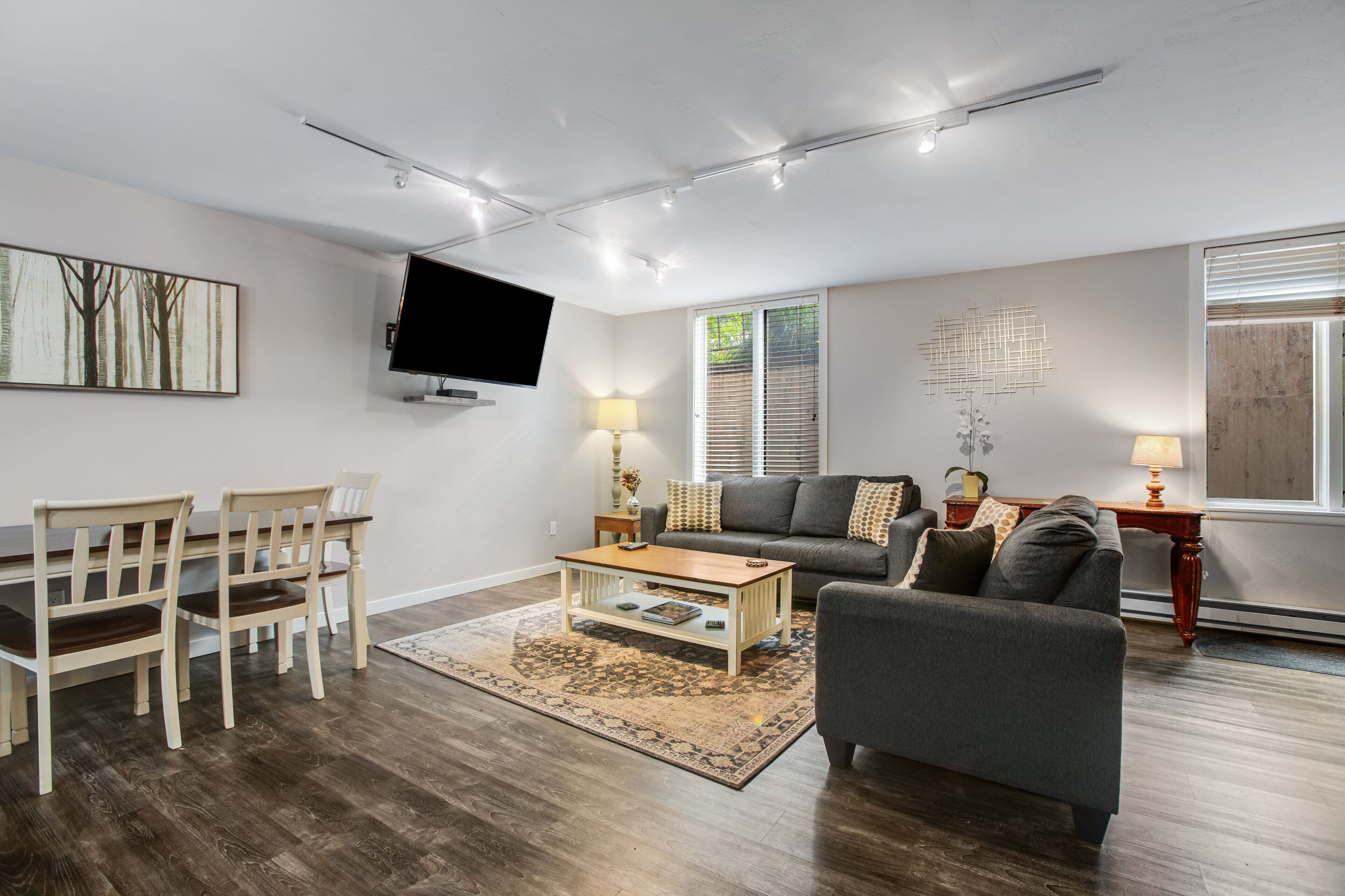 Ein modernes Wohnzimmer mit Flachbildfernseher, grauem Sofa, Couchtisch aus Holz und Essbereich mit Tisch und Stühlen.