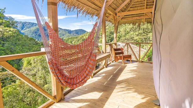 Una terraza de madera con una hamaca, bancos de madera y una estructura de techo de paja que da vista a un paisaje montañoso.