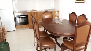 A dining area with a wooden table and chairs, a vase with dried plants, and a kitchen in the background.