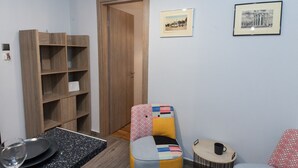 Een gezellig, compact ingerichte ruimte met een houten boekenkast, een kleine eettafel met borden en een glas, een kleurrijke fauteuil, een rond bijzettafeltje met een kopje, en twee ingelijste foto’s aan de muur.