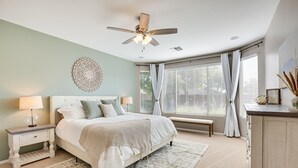 Une chambre à coucher comprenant un lit, des tables de chevet, un ventilateur de plafond, une fenêtre avec des rideaux et un banc.