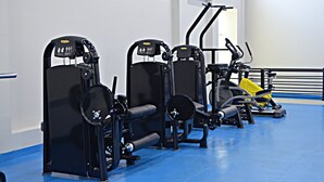 Une rangée d’appareils de musculation dans une salle de sport, avec un sol bleu et des murs blancs.