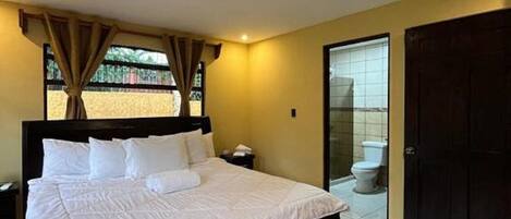 A bedroom with a large bed, a nightstand, a window with curtains, and a view of the outside.