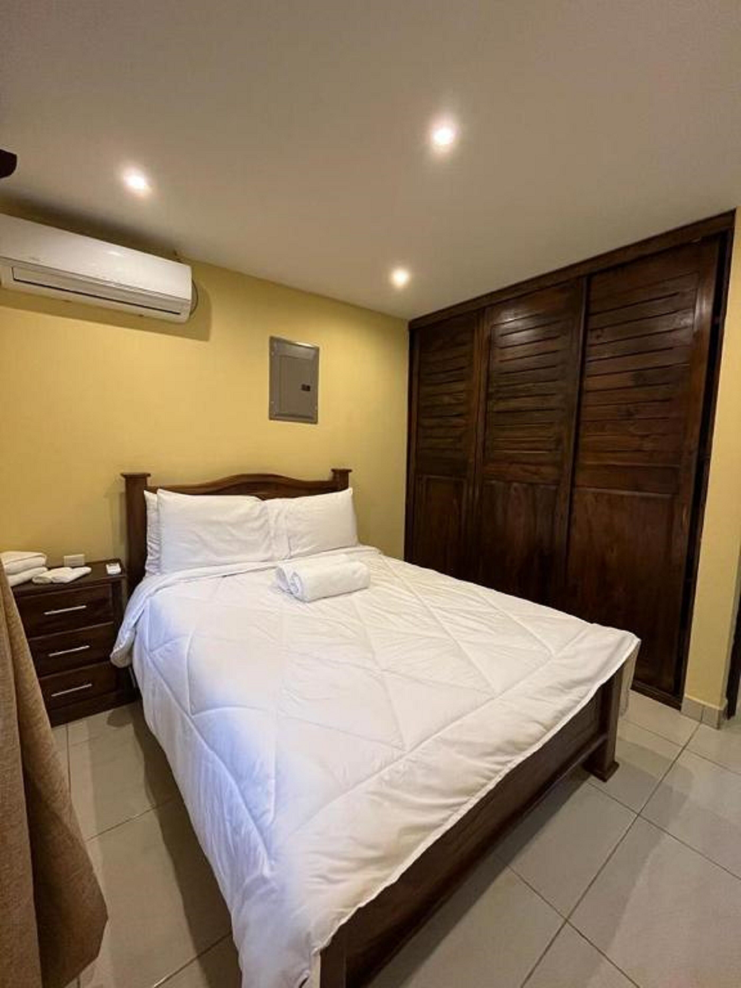 A bedroom with a bed, a nightstand, an air conditioning unit, a wooden wardrobe, and a wall-mounted electrical outlet.
