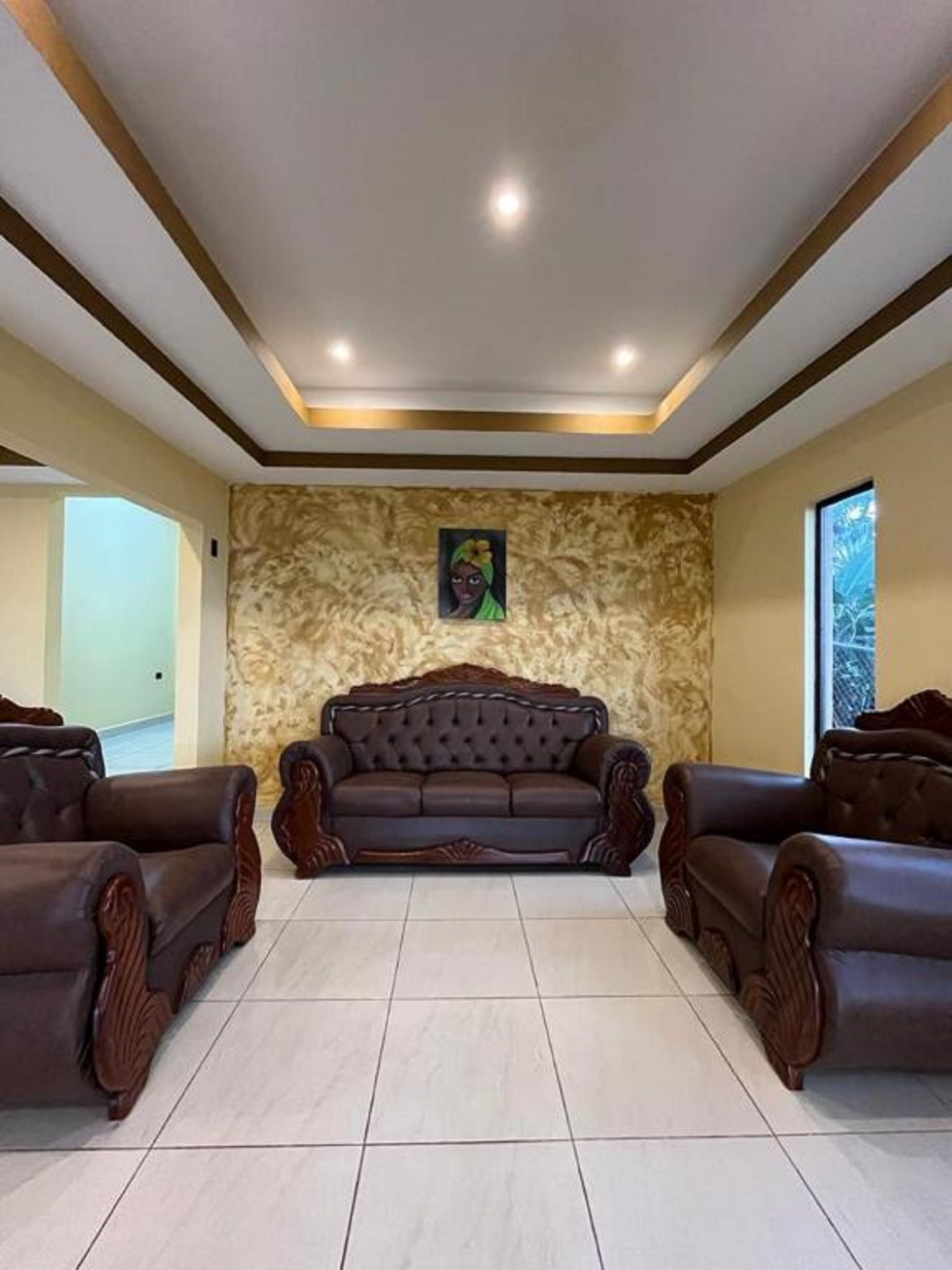 A spacious living room with a large, dark brown leather sofa centered on a tiled floor, flanked by two matching armchairs. The room features a high ceiling with recessed lighting and a textured wall with a framed artwork above the sofa.