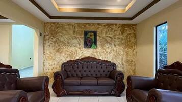 A spacious living room with a large, dark brown leather sofa centered on a tiled floor, flanked by two matching armchairs. The room features a high ceiling with recessed lighting and a textured wall with a framed artwork above the sofa.