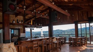 Um espaço de bar rústico com mobiliário de madeira, parede de pedra e vista para uma paisagem montanhosa.