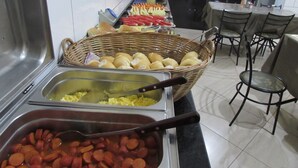 Een buffetgedeelte met diverse gerechten, waaronder een mand met broodjes, een bakje plakjes worst en een schaal roerei.