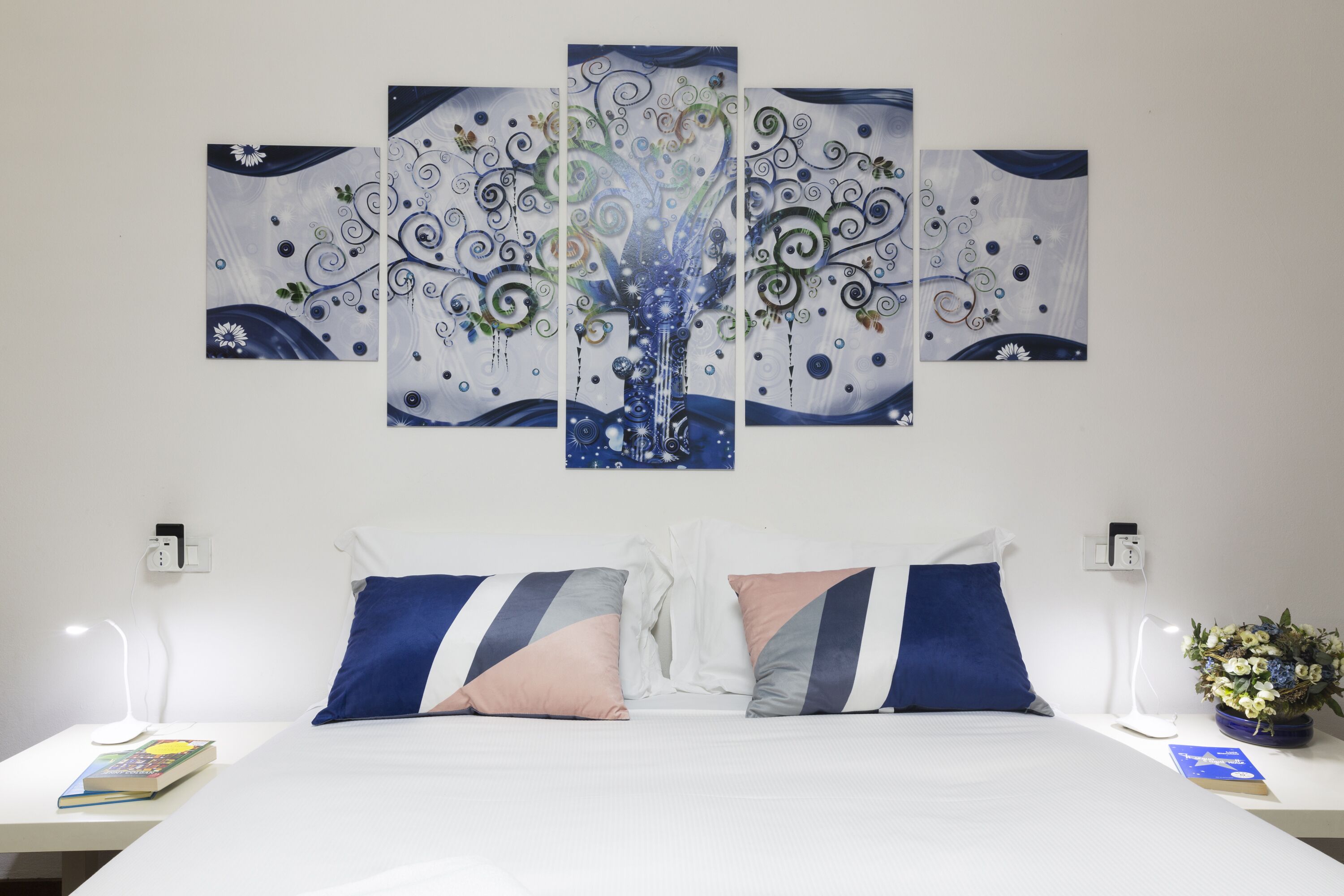 A hotel room with a bed, pillows, a nightstand, a lamp, a book, and a wall art piece.