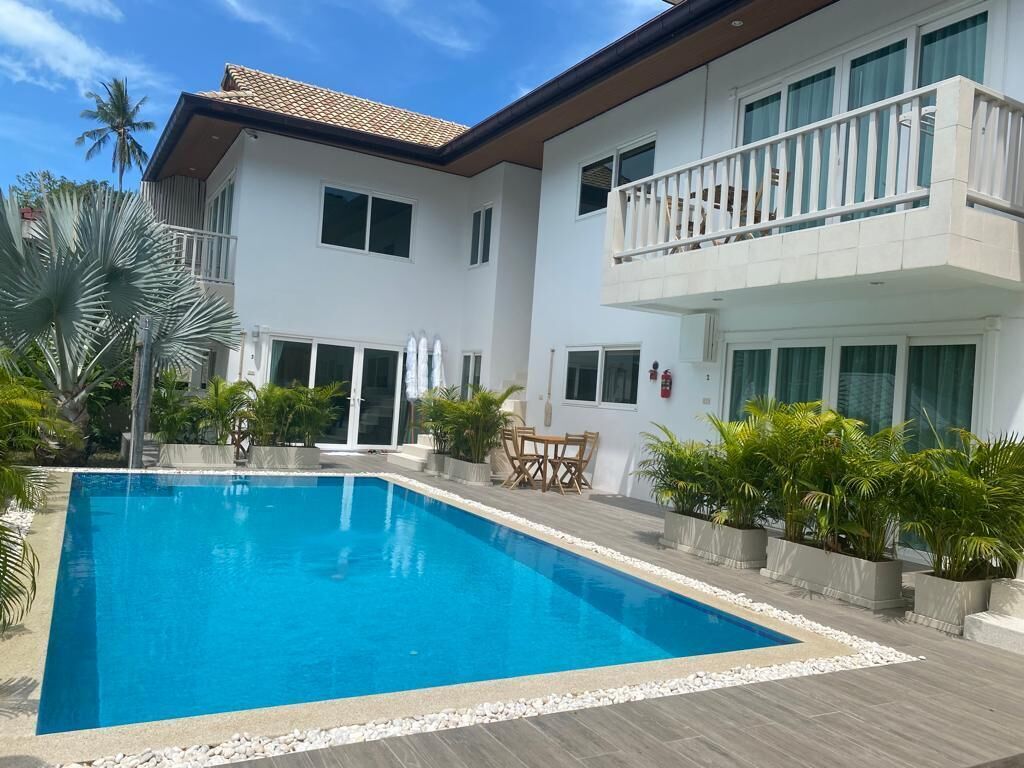 A modern two-story house with a swimming pool, surrounded by potted plants and palm trees.