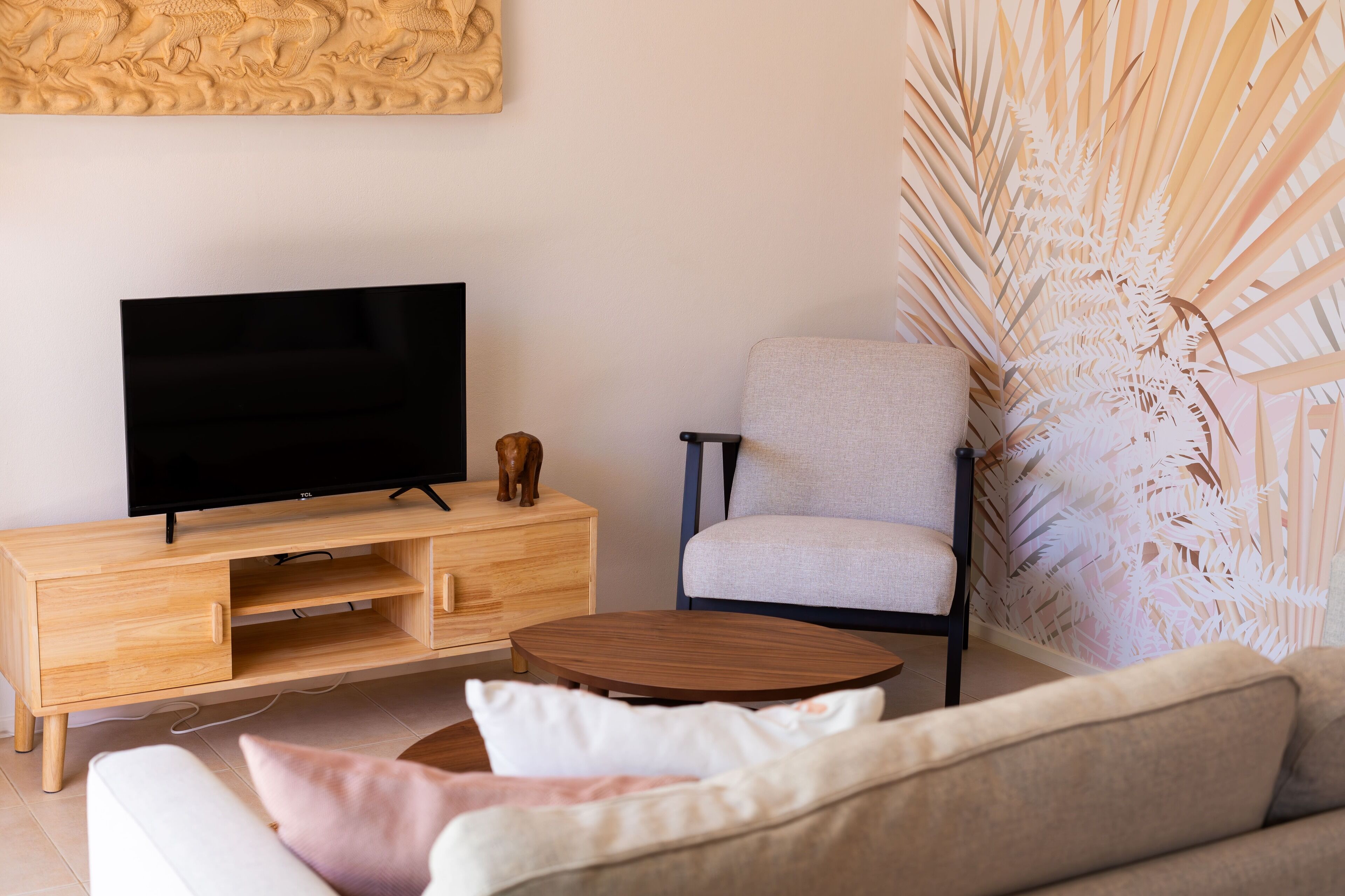 A modern living room with a flat-screen TV on a wooden TV stand, a light grey armchair, a round wooden coffee table, and a beige sofa with cushions.