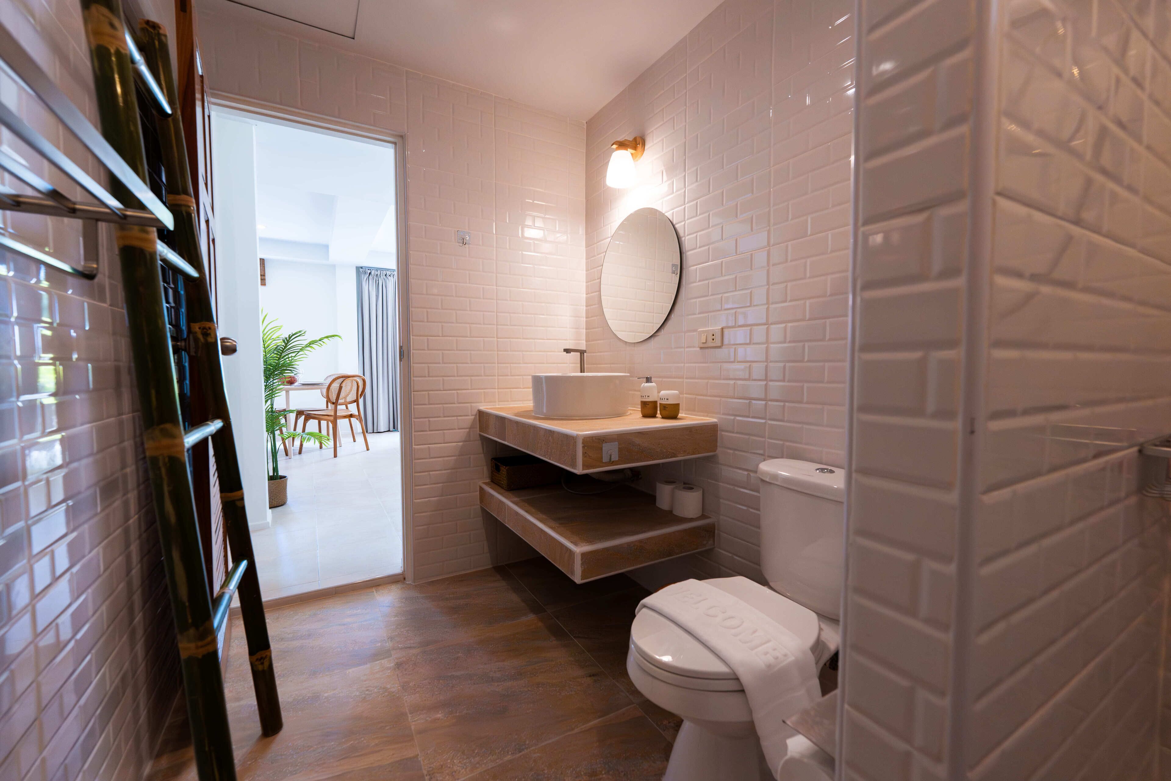 Un baño moderno con azulejos blancos tipo metro, un espejo redondo, un inodoro blanco y un mueble de tocador de madera.