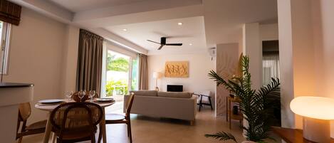 A spacious living area with a sofa, dining table, and chairs, a ceiling fan, and a large window with curtains.