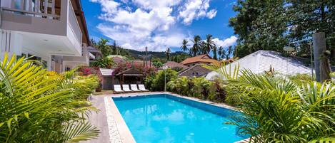 Piscina rodeada de plantas tropicales, una terraza de madera y vistas a una zona residencial con casas y palmeras.