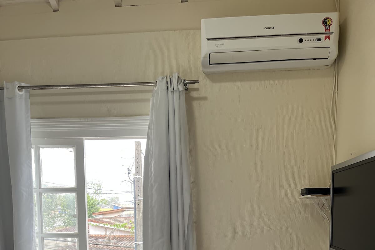 Ein Zimmer mit einem Bett, einer Klimaanlage, einem Fenster mit Vorhängen, einem Fernseher und einem kleinen Tisch mit Bilderrahmen und Fernbedienung.