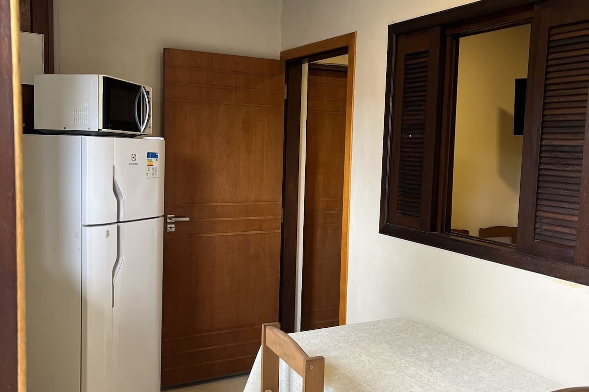 Eine kleine Küche mit Kühlschrank, Mikrowelle und einer Holztür. Ein Tisch mit weißem Tischläufer und zwei Stühle sind vorhanden.