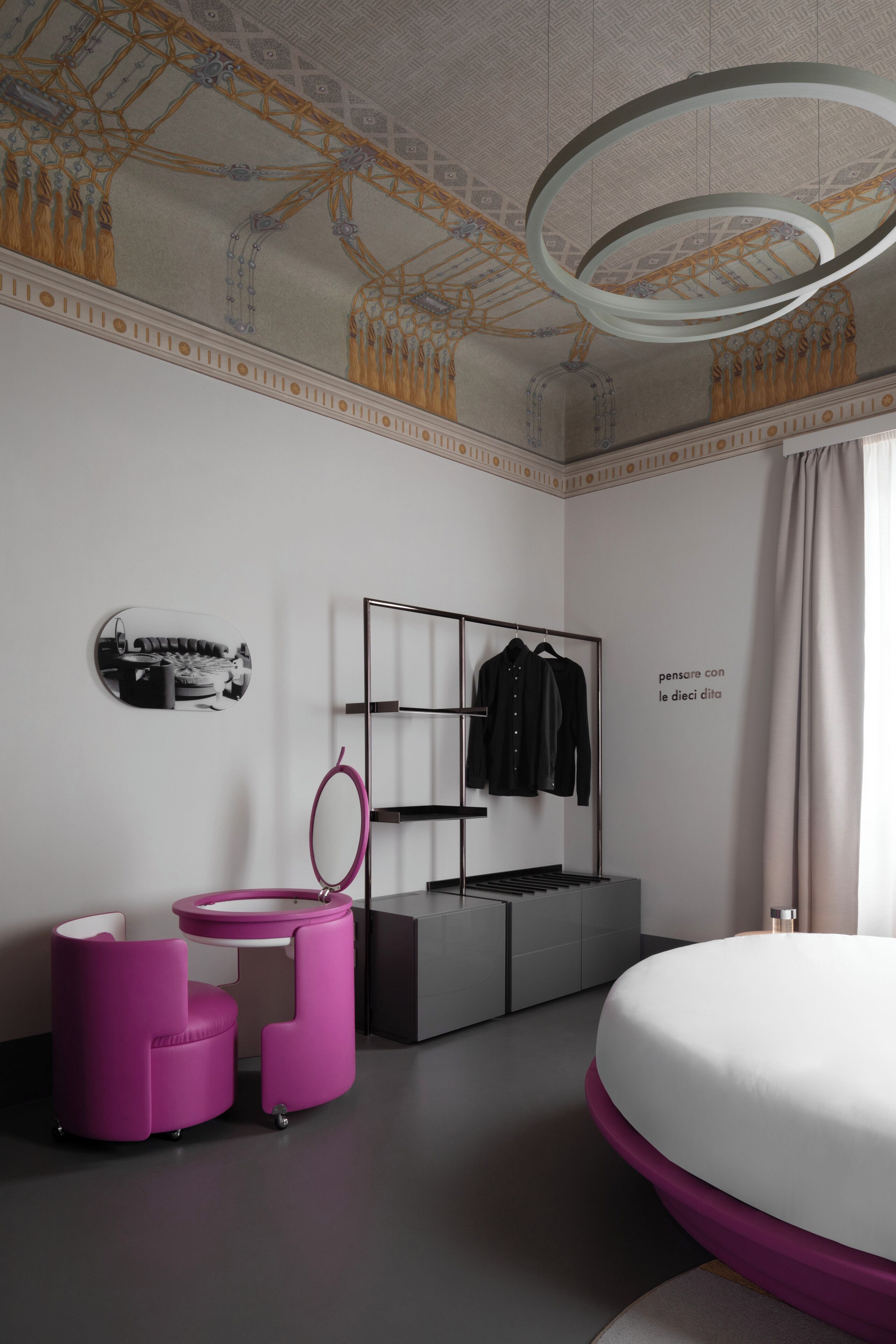 A modern bedroom with a bed, a vanity with a mirror, a pink chair, a black coat rack, and a decorative ceiling.