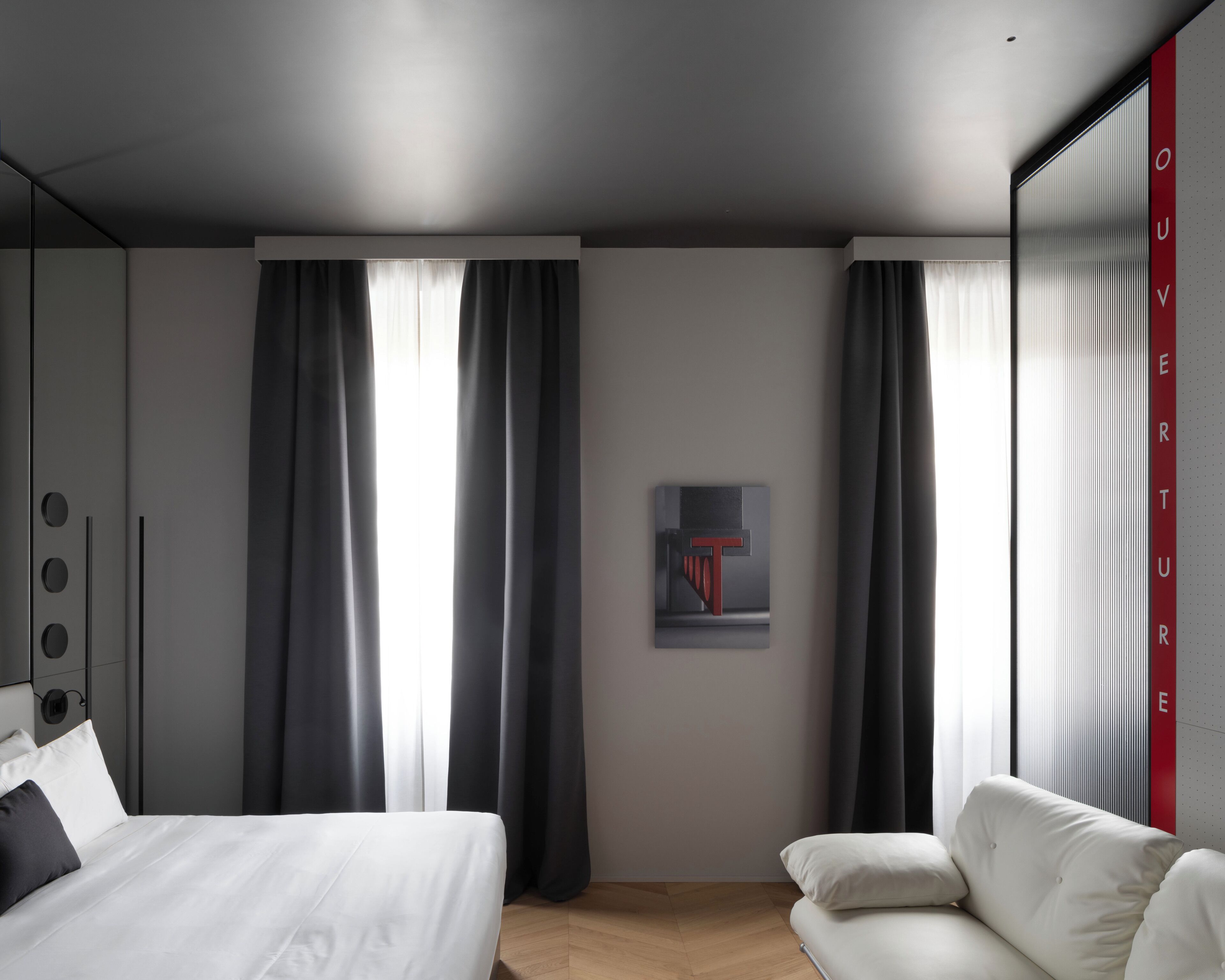 Quarto de hotel moderno com cama, sofá e uma grande janela com cortinas.