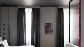 Quarto de hotel moderno com cama, sofá e uma grande janela com cortinas.