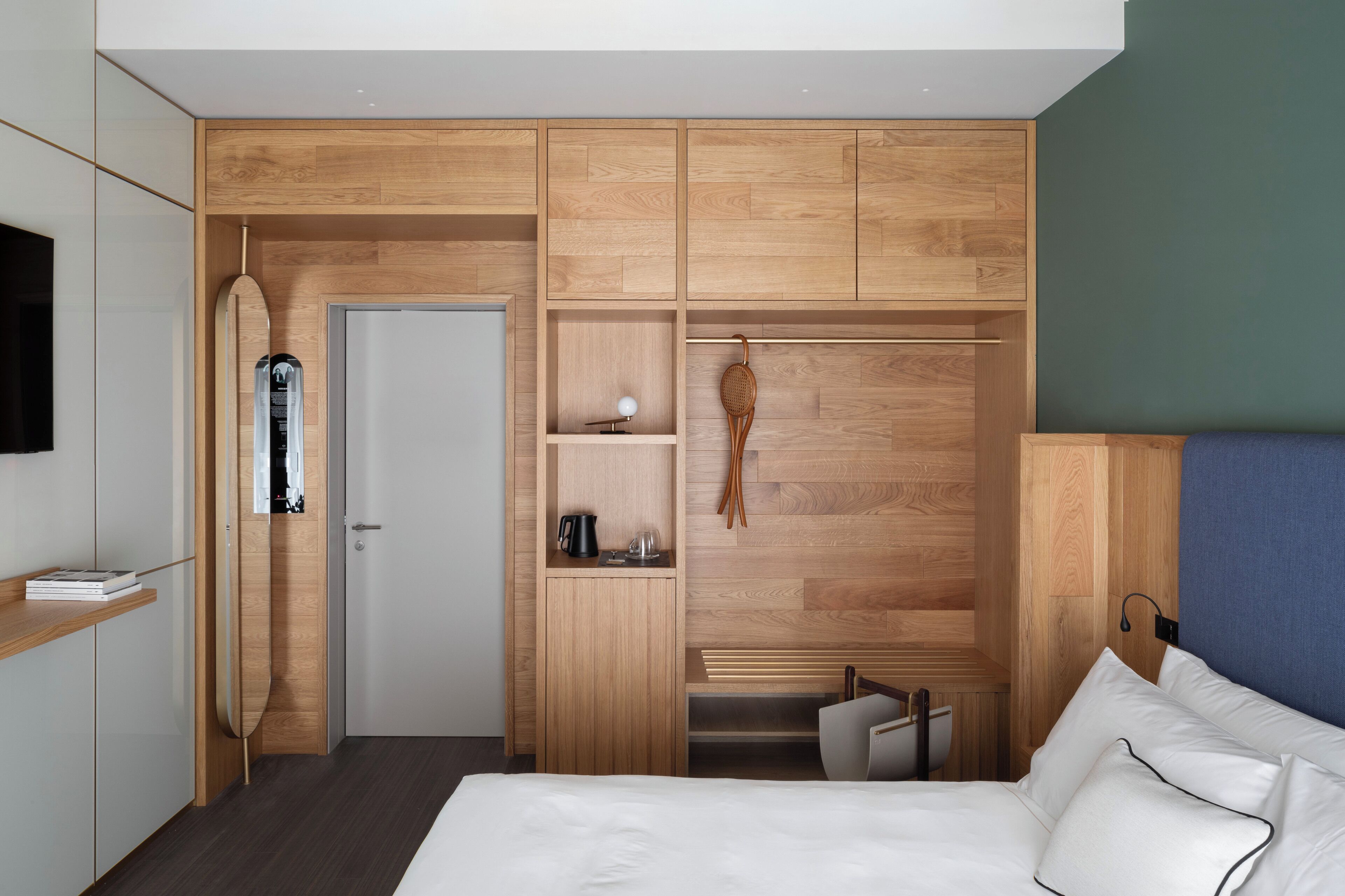 A modern hotel room with a bed, a wardrobe with multiple compartments, a small seating area, and a television mounted on the wall.