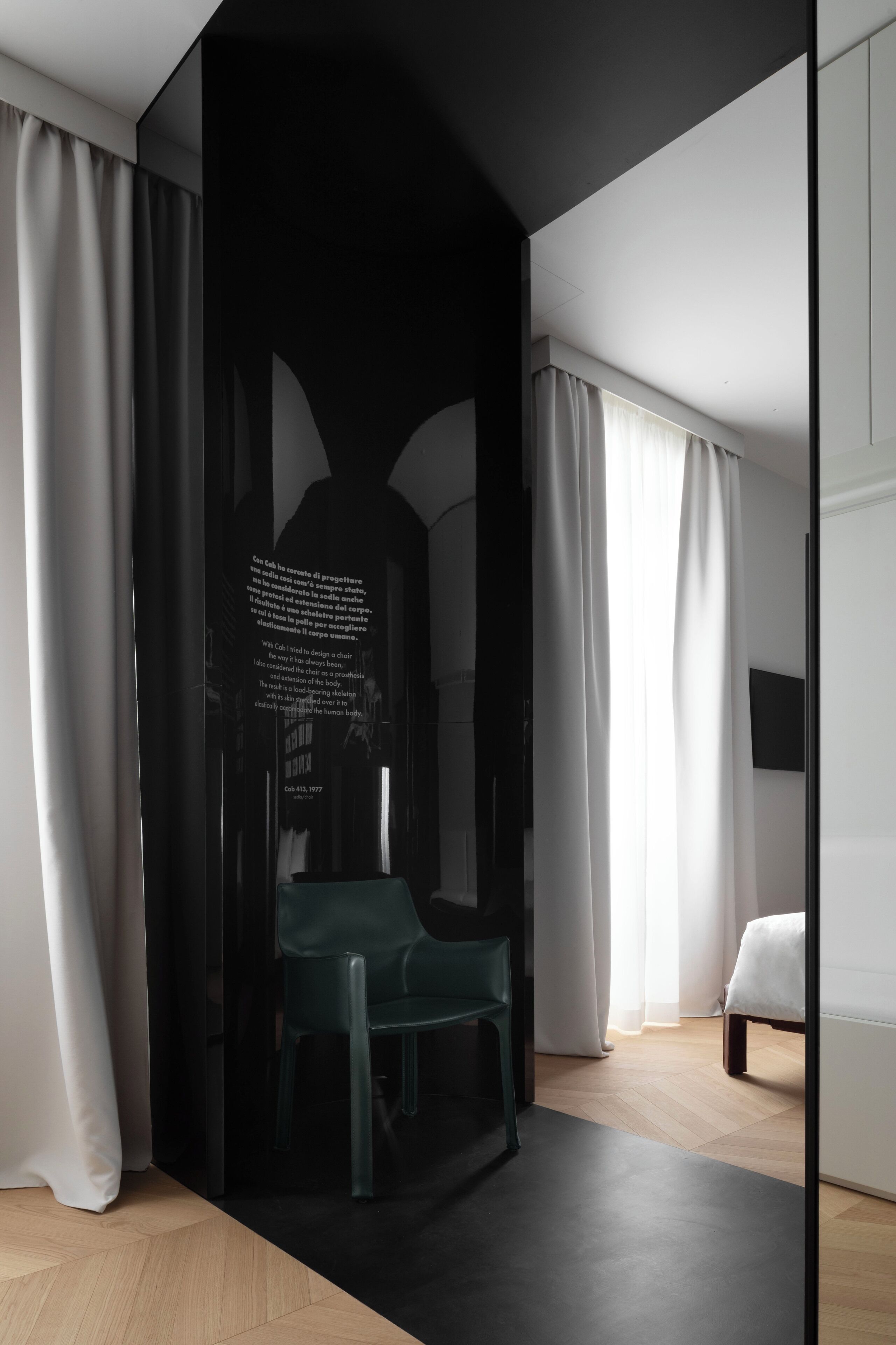 A modern hotel room with a dark accent wall, a green chair, a bed, and a large window with curtains.