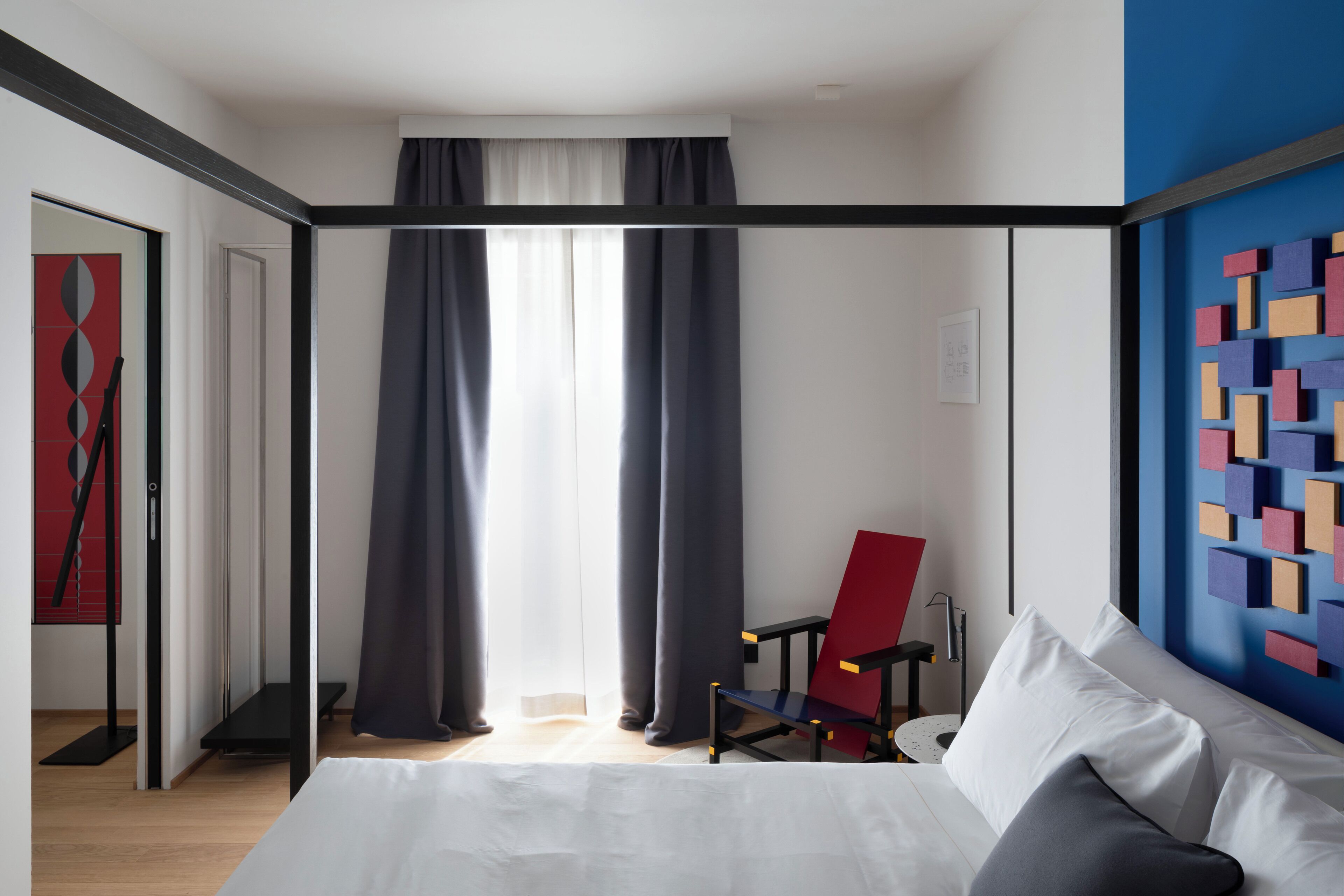 A hotel room with a bed, a chair, a desk, and a wall with colorful geometric decorations.