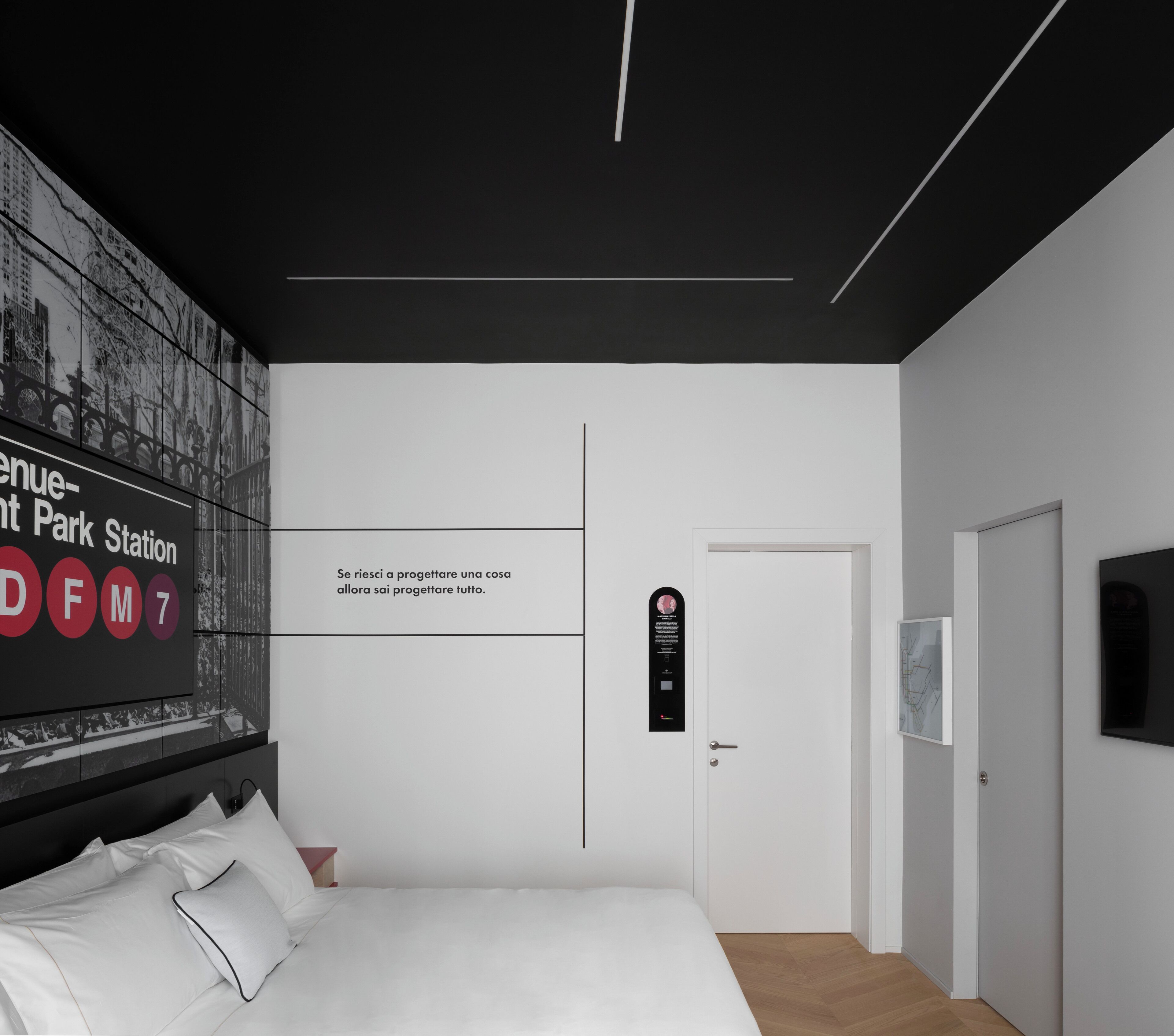 Habitació d'hotel moderna amb un llit gran, un televisor fixat a la paret que mostra un paisatge urbà, una porta amb panell de seguretat i un disseny minimalista.