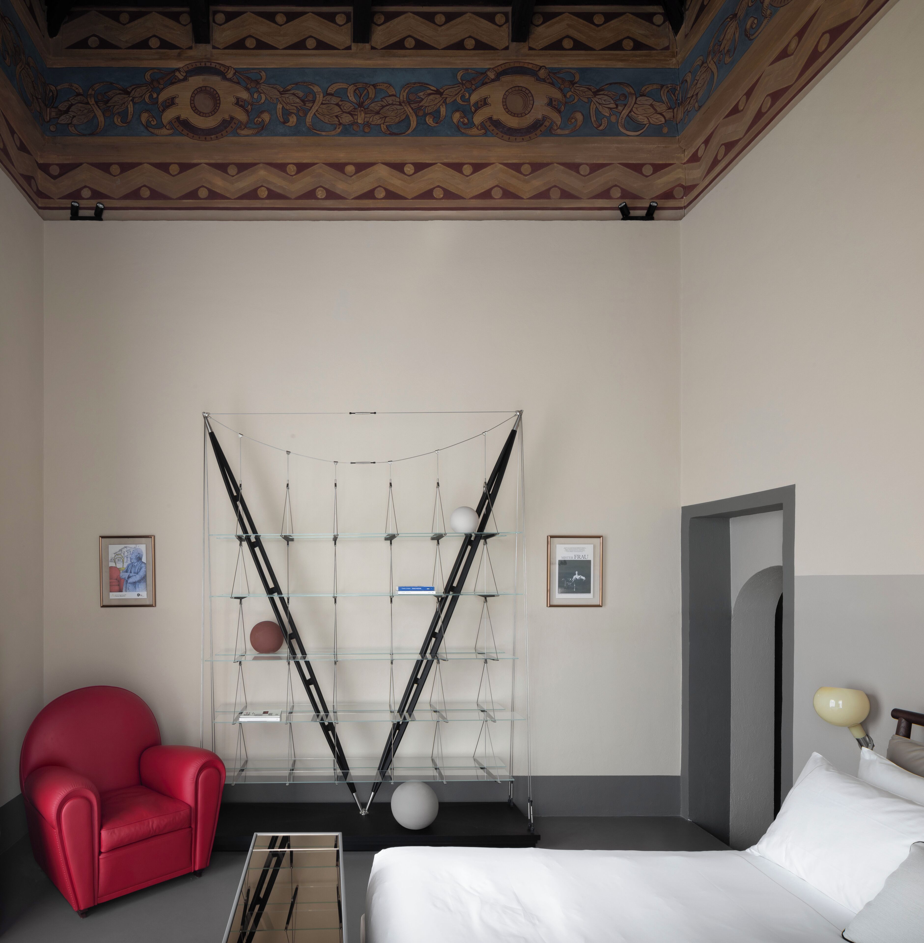 Une chambre d’hôtel avec un lit, un fauteuil rouge, un plafond décoratif et deux tableaux encadrés accrochés au mur.