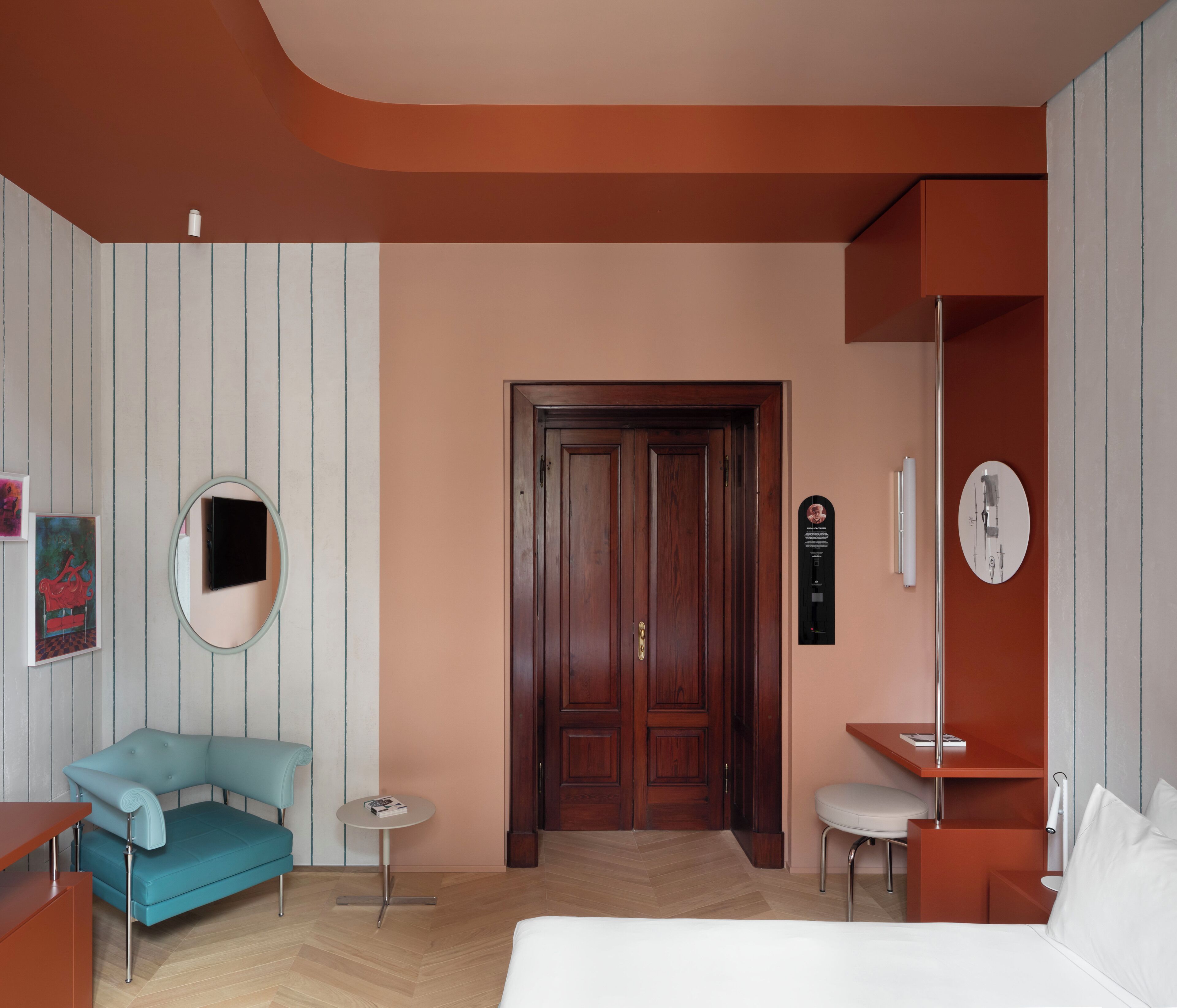 A hotel room with a wooden door, a round mirror, a blue armchair, a small round table, a white chair, and a bed with white bedding.