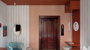 A hotel room with a wooden door, a round mirror, a blue armchair, a small round table, a white chair, and a bed with white bedding.
