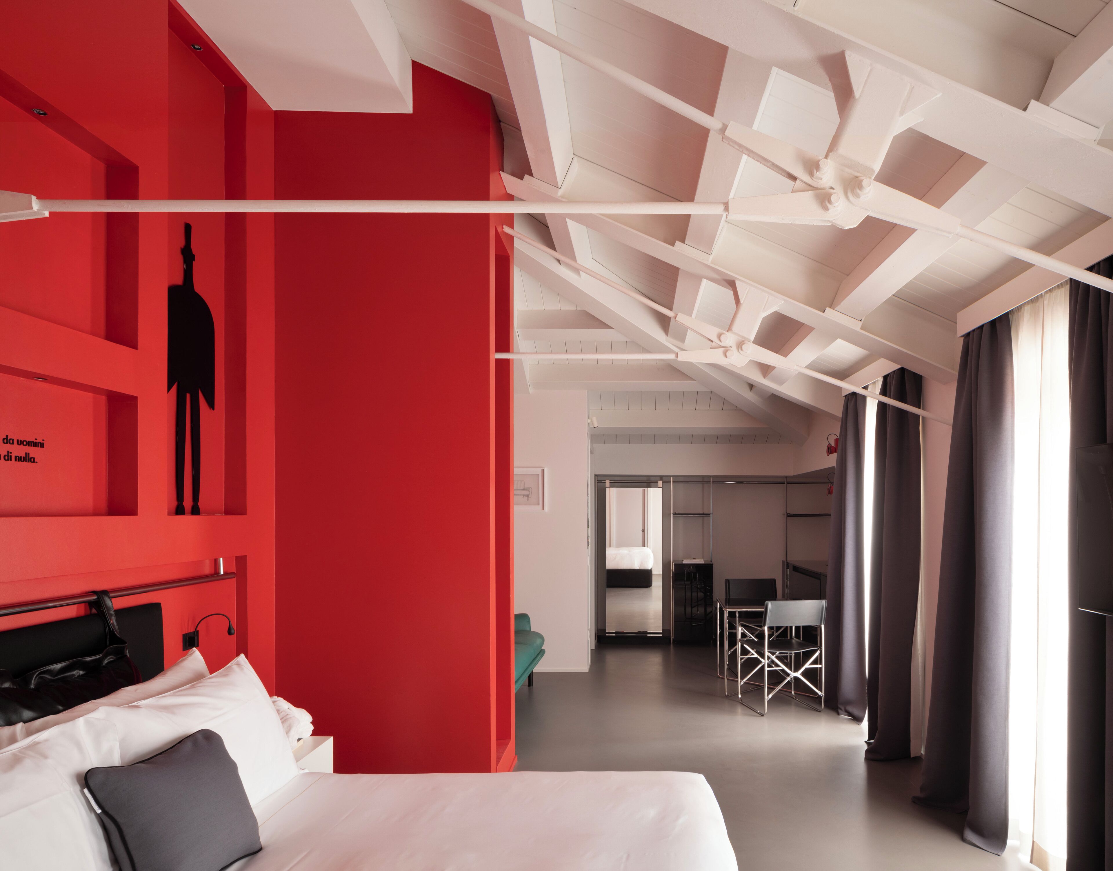 A modern hotel room with a red accent wall, a bed with white linens, a desk with a chair, and a view of the hallway.