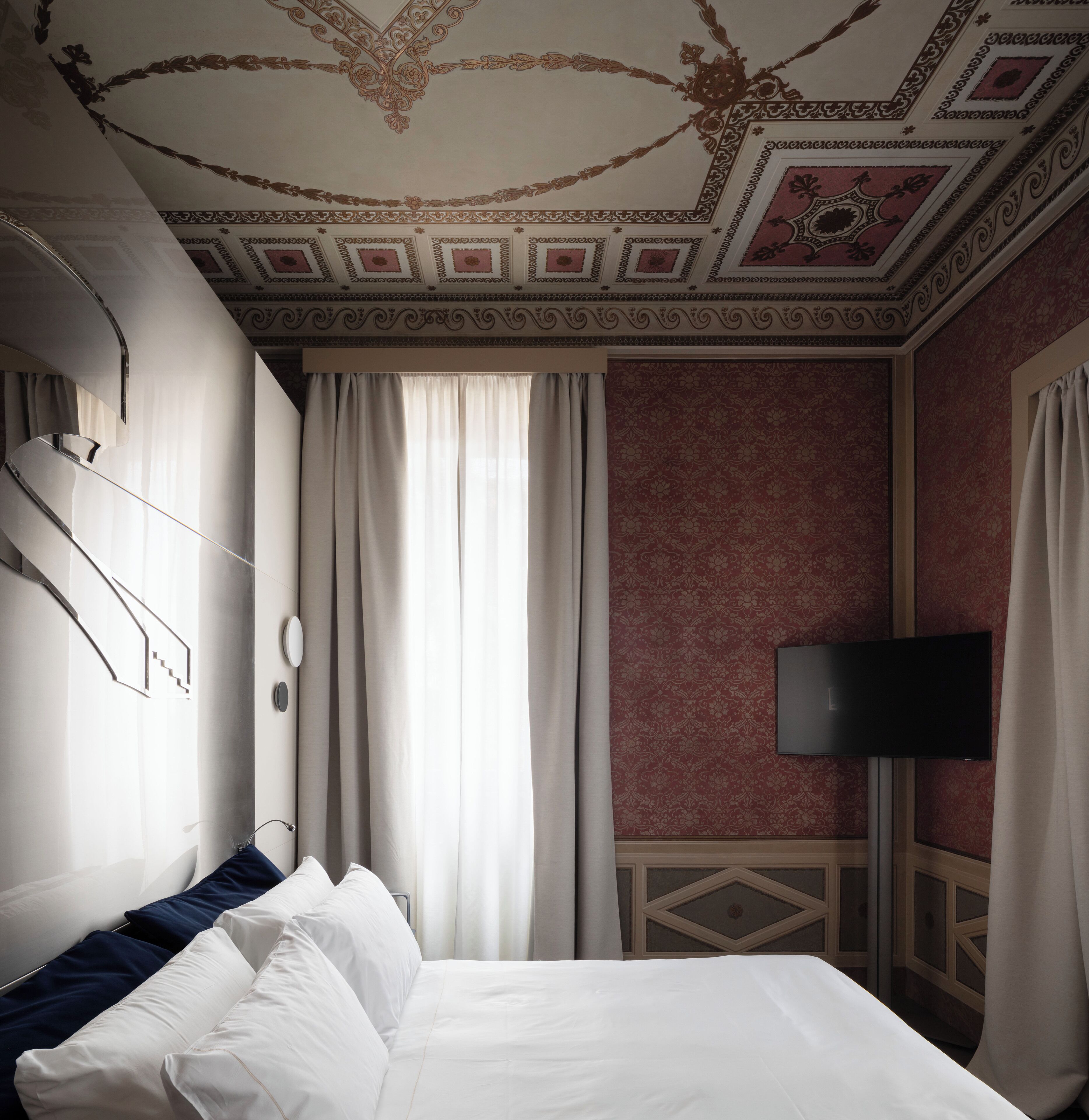 A hotel room with a bed, curtains, a flat-screen TV, and ornate ceiling design.
