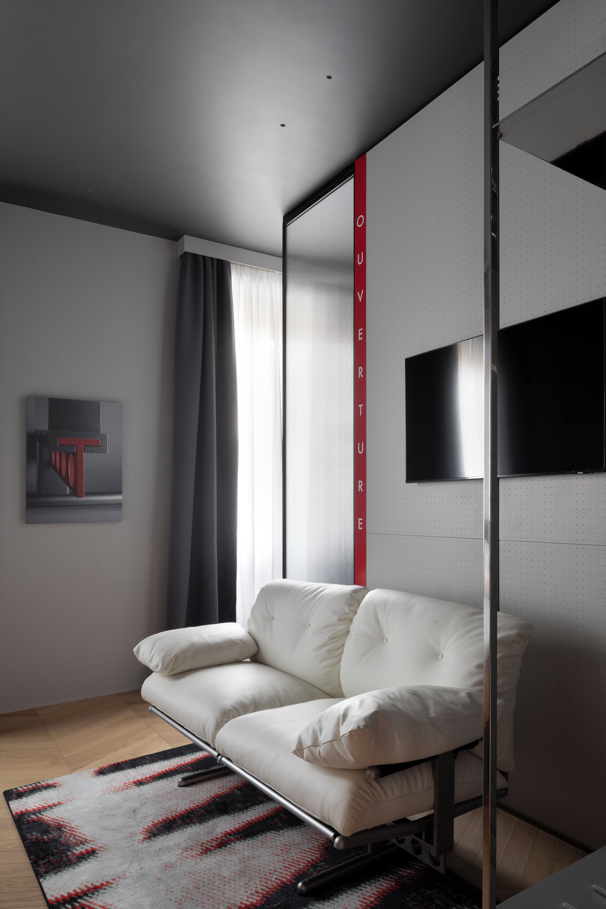 Ein modernes Hotelzimmer mit einem weißen Ledersofa, einem großen Fenster mit Vorhängen, einem an der Wand befestigten Flachbildfernseher und einem gemusterten Teppich.