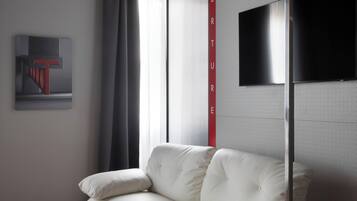Ein modernes Hotelzimmer mit einem weißen Ledersofa, einem großen Fenster mit Vorhängen, einem an der Wand befestigten Flachbildfernseher und einem gemusterten Teppich.